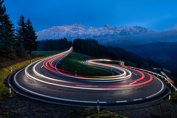Light Trail Of Moving Cars Driving Up A Panoramic Serpentine Road In The Swiss Alps - obrazy, fototapety, plakaty