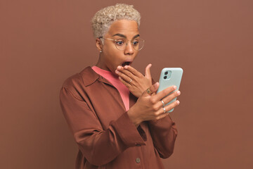 Young dazed ethnic African American woman holds phone and covers open mouth with hand indignant at news or inaccurate statement on Internet site stands on brown background. Surprise, indignation