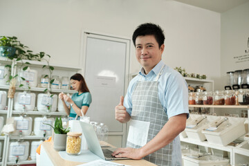 Portrait of happy Asian male shopkeeper smiles looks at camera, and thumbs up at refill store, natural products, zero-waste grocery, and plastic-free, eco environment-friendly, sustainable lifestyles.