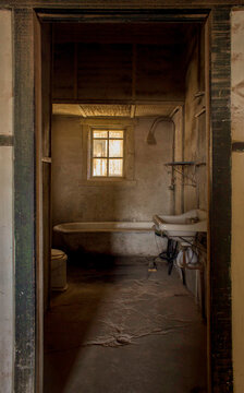 Antiguo ba&ntilde;o abandonado con una tina ducha, lavamanos de loza y una peque&ntilde;a ventana con reja. Luz tenue y sombras agradables