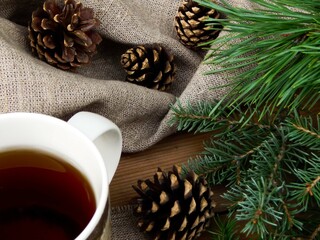 A mug standing on a linen napkin next to spruce branches and scattered cones, in the background a linen tablecloth, on a wooden table