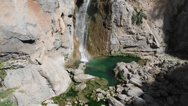 Still steady shot slowly zooming out as freshwater drops from a cliff at a bottom of a waterfall, forming a pond by carving a lake in the rock, an exotic green pond.