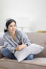 Obraz premium Photo of modern asian woman wearing headphones holding cell phone while lying on sofa in bright apartment