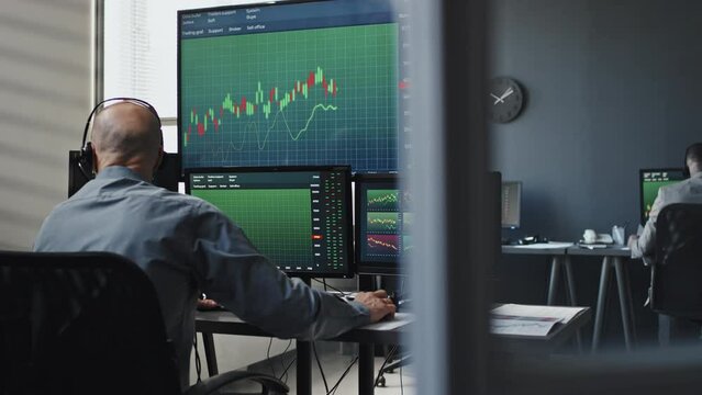 Rear view of professional stock trader wearing headphones working with graphs and stats on computer monitors and discussing something with client