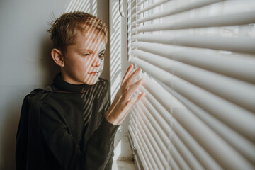 A boy looks out the window through the blinds waiting for friends to celebrate halloween