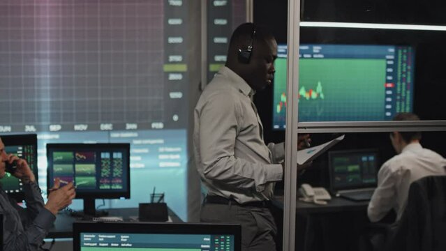 Young Black stock trading advisor wearing headset holding paper with graphs and charts talking on phone with client late in evening, his Caucasian colleagues working on background