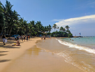 Hikkaduwa, Sri Lanka - March 6, 2022: Beautiful view of the beach in Hikkaduwa with green palm trees. People relax, sunbathe and swim in the Indian Ocean