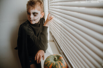 A boy looks out the window through the blinds waiting for friends to celebrate halloween