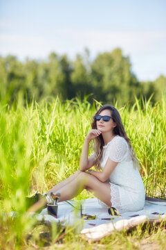 Young blonde woman in white dress and sunglasses is sitting on a blanket in tall grass.