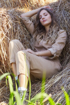 Young blonde woman in a beige jumpsuit is sleeping on the rolls of hay.