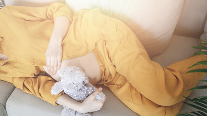Pregnancy. Young pregnant woman wearing maternity clothes relaxing on home sofa with teddy bear baby toy. Pain of contractions during childbirth. Child expecting. Family planning. Natural pregnancy