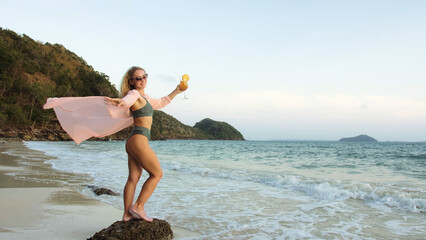 Attractive woman stand on a reef rock stone in sea on golden sunset. Girl on tropical beach in green swimsuit and flutter in wind pink tunic silk shirt cape, drinks her orange cocktail Pina Colada