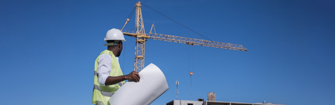 African American architect compares building construction to drawings of future project. Black constructor controls building at back sunlight