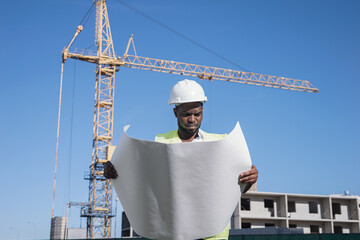 African American architect compares building construction to drawings of future project. Black constructor controls building at back sunlight