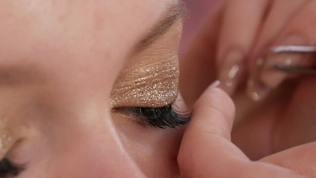 Make-up artist sticks long black false eyelashes on the eyes of a young beautiful girl model with bright evening gold makeup close up view