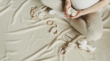 Baby shower, infant care concept. Pregnant woman holding wooden newborn stuff, baby accessories - shoes, toys on beige background. Local eco children products, baby shower. Top view, copy space
