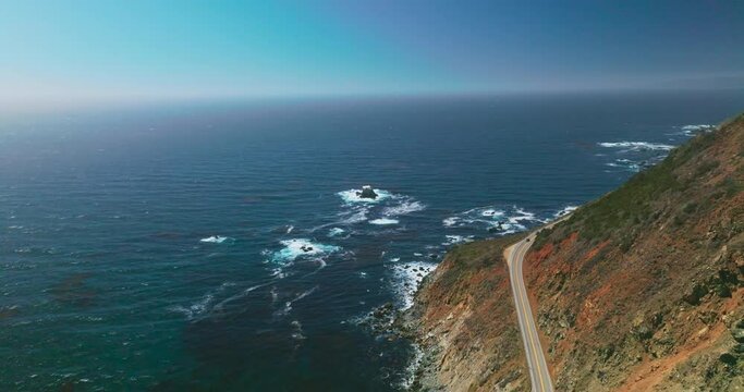 Rocky shore with highway over cliffs. Beautiful endless blue Pacific Ocean interflowing with clear azure sky. Aerial view.