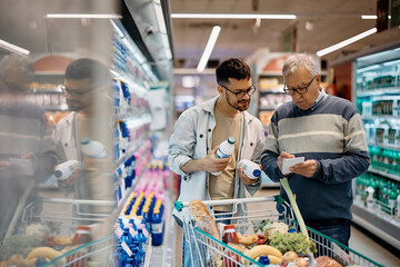 Young man and his senior father going through shopping list while buying in supermarket. - obrazy, fototapety, plakaty