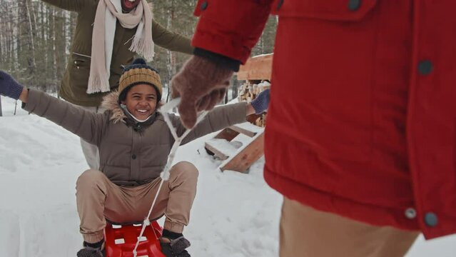 Slow motion of happy African American boy enjoying sledging on winter day, his father pulling the sledge and mother following them