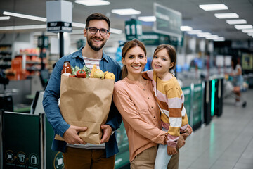 Happy parents and their small daughter buying in supermarket and looking at camera.