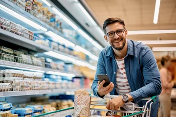 Foto op Plexiglas Happy man using mobile phone app while buying groceries in supermarket and looking at camera. © Drazen