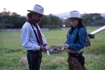 Cowboy man standing at the green land near mountain side teaching cow girl how to handle revolver...