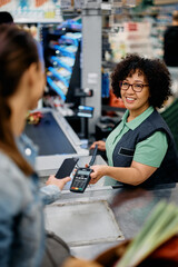 Happy cashier holds credit card reader while customer is paying with smart phone at supermarket.