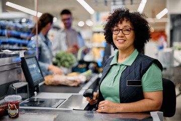 Happy woman working as cashier at supermarket checkout and looking at camera. - obrazy, fototapety, plakaty