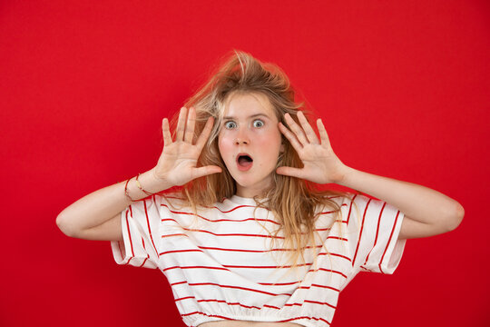 Open eyed young lady stand with raised hands with spread fingers near face on empty red background. Nice girl with wild hair have fun and show surprise emotion. Astonishment, wow effect