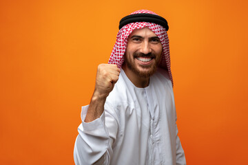 Successful smiling young Arab man celebrating victory on orange background