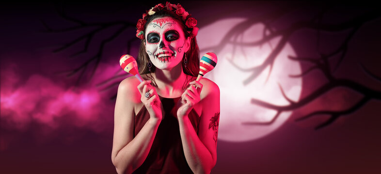 Young woman with painted skull on her face and maracas at night. Celebration of Mexico's Day of the Dead (El Dia de Muertos)