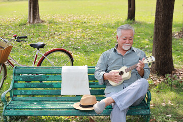 Happy smiling asian senior man with beard sitting on bench playing ukulele and singing a song in...