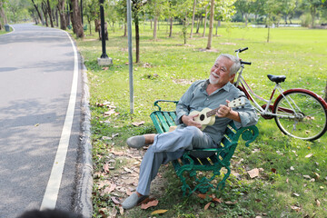Happy smiling asian senior man with beard sitting on bench playing ukulele and singing a song in garden park outdoor. Musical and relaxation makes elder male happiness. Health care lifestyle concept.