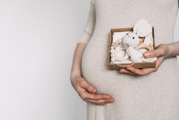 Pregnant woman holding gift, wooden newborn stuff, baby accessories, teether, toys on beige background. Pregnancy, maternity, preparation, baby expectation concept. Baby shower, infant care concept