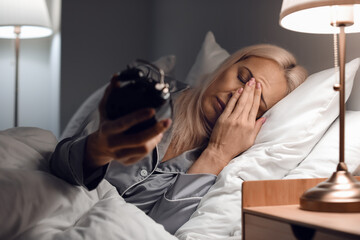 Stressed mature woman with alarm clock lying in bed at night - obrazy, fototapety, plakaty