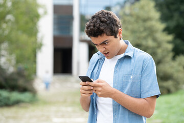 Fototapeta premium Disappointed hispanic boy looking at phone with nasty face. Student receiving bad news on internet.