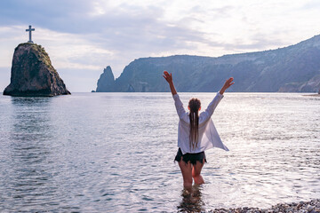The girl stands on the shore and looks at the sea. Her hands are raised up. She wears a white shirt and her hair is in a braid.