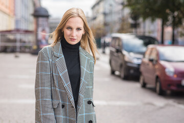Fototapeta premium Portrait of a young and elegant Caucasian blonde girl in autumn in the city. The concept of femininity and style. A pretty girl is happy to pose in the city on the street in a coat.