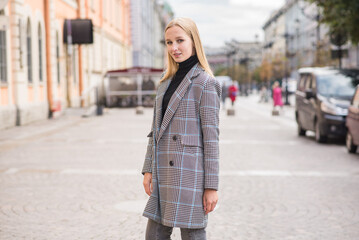 Fototapeta premium Portrait of a young and elegant Caucasian blonde girl in autumn in the city. The concept of femininity and style. A pretty girl is happy to pose in the city on the street in a coat.