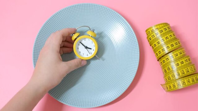 Female hand holding an alarm clock on background of an empty plate, concept of intermittent fasting.