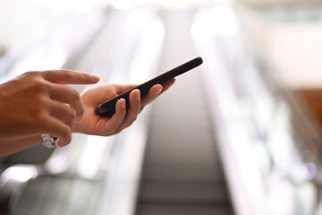 Woman hand typing on smartphone with blurred moving escalator and sunlight in background