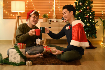 Two happy man next to a decorated Christmas tree and enjoying drinking hot chocolate together