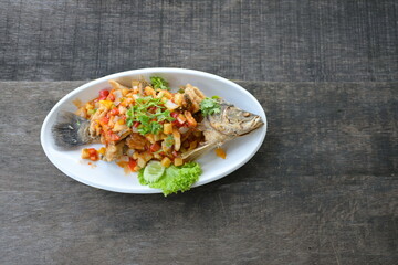 Deep-Fried snapper with Three-Flavoured Sauce. Deep Fried Fish with Chili Sauce, Pineapple, onion, tomato, carrot (all diced) topped with shredded coriander served in a white plate on a wood table.
