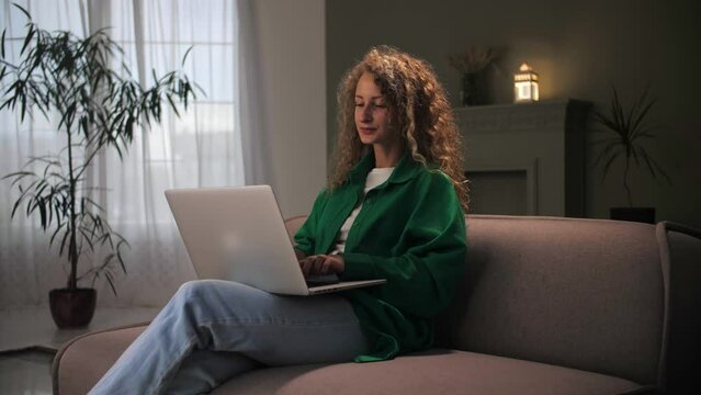 Woman work remotely using laptop sitting at couch.Curly hair girl looking at computer typing on keyboard in home interior.Female browsing internet on notebook surfing web online shopping concept.