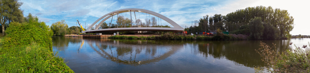panoramic view on the preapering of a bridge on a construction site  with crane and construction material on a riverside surrounded by trees with the reflection of the bridge in the water of the river