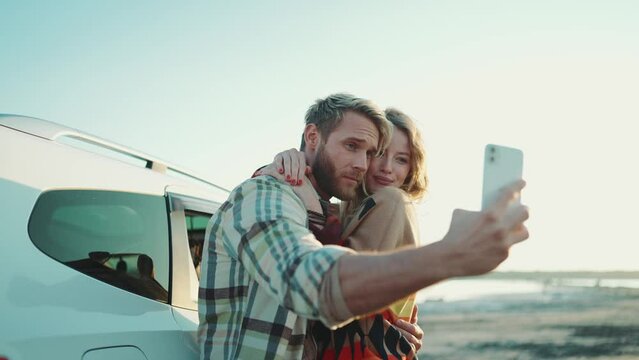 Positive blond couple making selfie photo on mobile near car outdoors