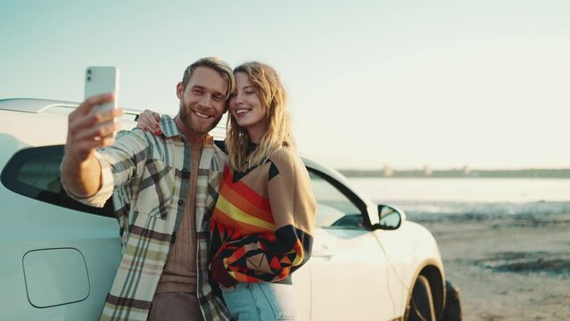 Happy blond couple making selfie photo on mobile near car outdoors