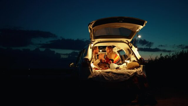 Happy blond young couple holding hands and talking while sitting in the trunk of the car 
