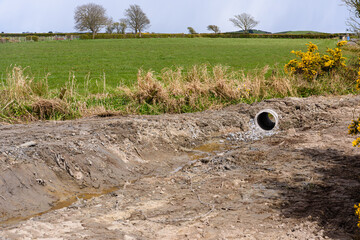 A drainage ditch is dug in a field, with a small culvert pipe to drain water into a small stream. - obrazy, fototapety, plakaty