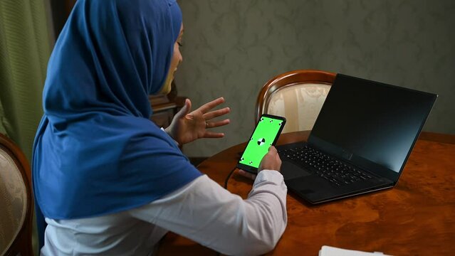 Rear view of a Muslim woman in hijab, talking during an online conference, using her modern smartphone with green chromakey screen with copy advertising space. Ideal video for insert promotional clip